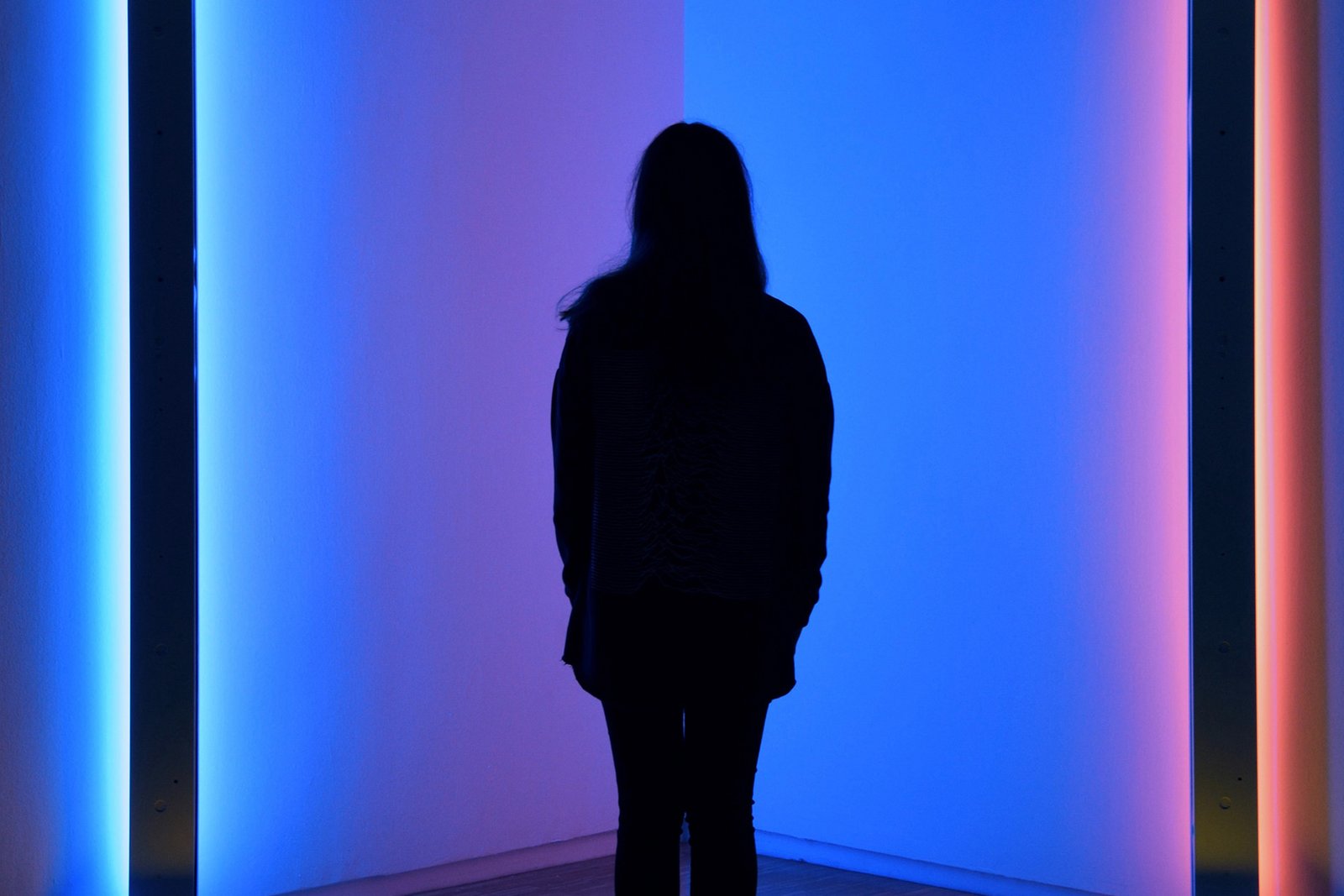 Im Halbdunkel zeigt sich die schwarze Silhouette einer jungen Frau in einer Ecke zwischen einem blauen und einem roten Lichtbündel.