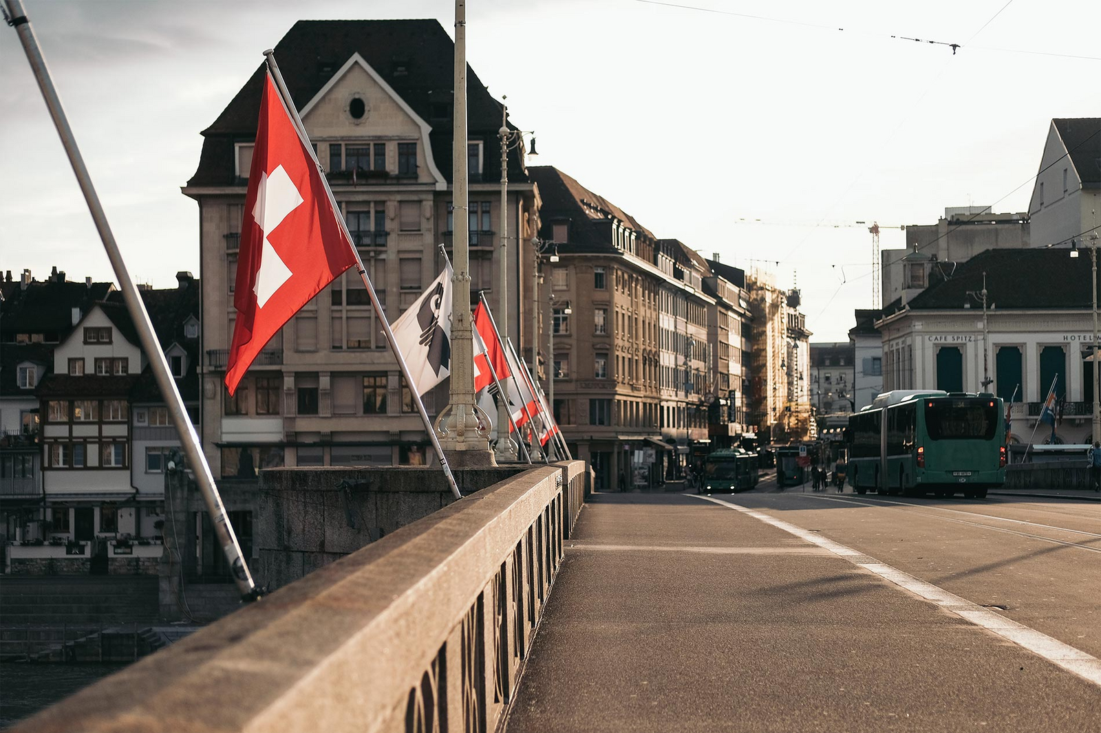 Des drapeaux suisses et du canton de Bâle-Ville flottent au vent depuis un pont à Bâle. En arrière-plan, un bus s'engouffre dans une rue surplombée par des immeubles anciens.