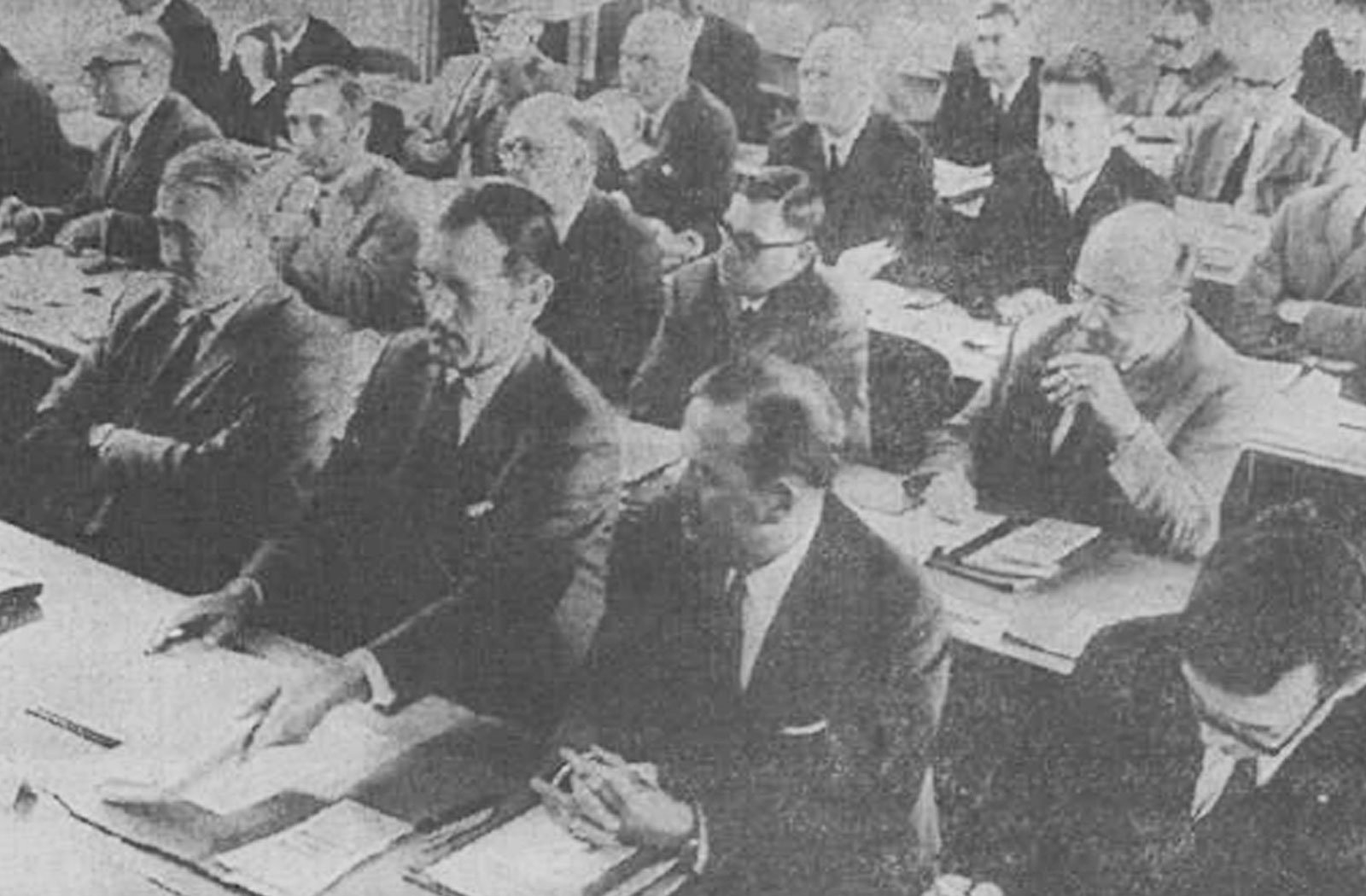Una ventina di uomini in giacca e cravatta ascoltano seduti una conferenza e prendono appunti.