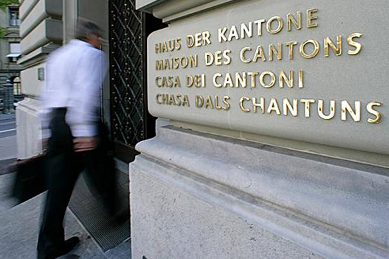In primo piano l'insegna della Casa dei Cantoni tradotta nelle quattro lingue nazionali; sullo sfondo un uomo entra nell’edificio. 