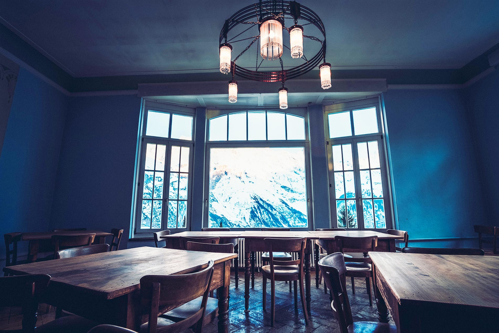 Une grande fenêtre avec vue sur les Alpes enneigées éclaire une volumineuse salle à manger meublée de tables et de chaises en bois massif.